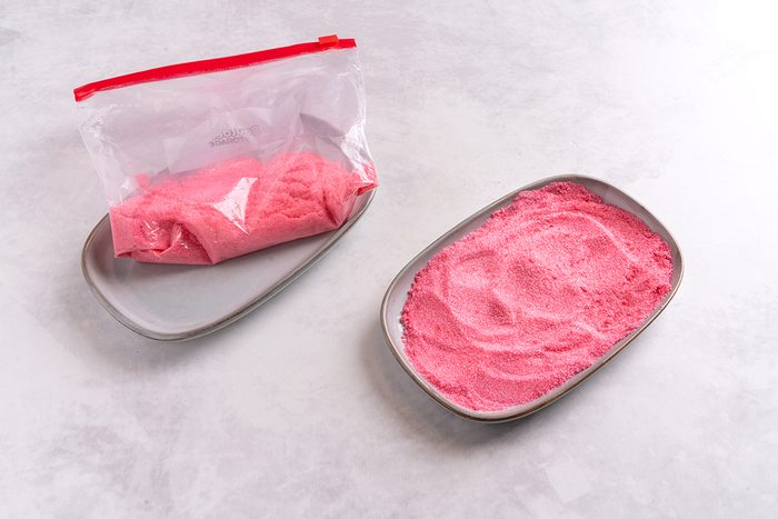 A sealed plastic bag filled with pink powder lies next to an open metal tray containing more of the same pink powder. Both are placed on a light gray, textured surface.