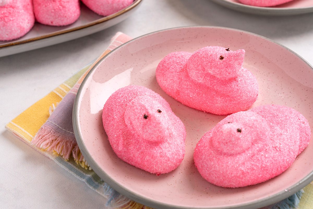Plate with three pink marshmallow chicks, resembling chicks or ducks, on a table. The plate is on top of colorful napkins.