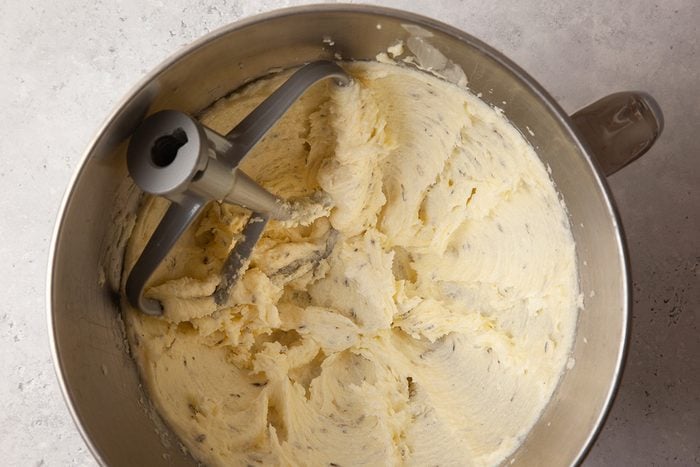 A metal mixing bowl filled with creamy, light beige dough or batter, with a paddle attachment. The mixture is smooth and evenly spread around the bowl. The bowl is set on a light-colored countertop.