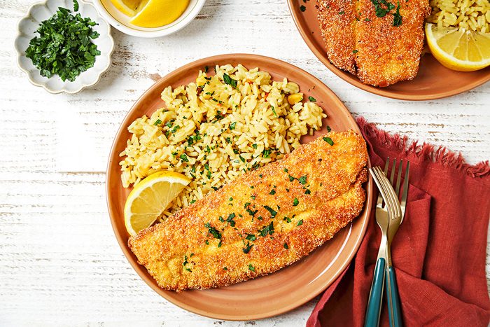 A plate with breaded fish fillet topped with chopped parsley, rice pilaf, and a lemon wedge on a light wooden surface. Next to it are a small dish of parsley, a red cloth napkin, and a fork and knife. A second plate is partially visible.