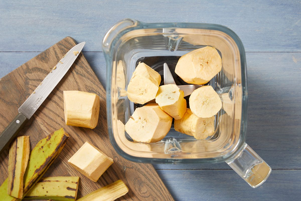 Sliced plantains in a blender jar, with additional unpeeled plantain slices and a knife on a wooden cutting board. The background is a light blue surface.