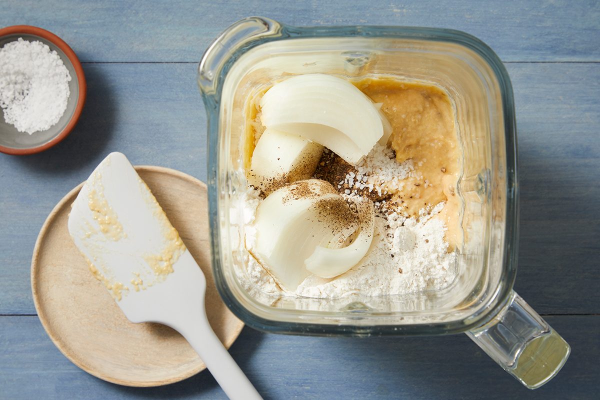 A blender filled with ingredients, including onion slices and spices, sits on a blue surface. Next to it are a spatula with some residue on a round plate and a small bowl of salt.