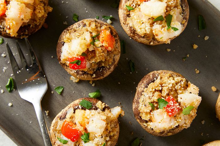 Stuffed mushrooms filled with a mixture of breadcrumbs, diced red peppers, and herbs, are arranged on a gray plate. A fork and scattered parsley leaves add to the presentation.