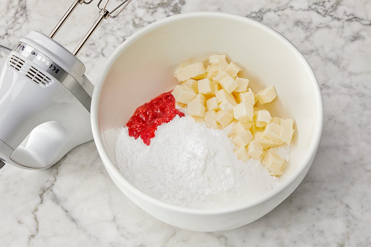 A mixing bowl on a marble countertop contains cubed butter, powdered sugar, and red-colored paste. An electric hand mixer is placed nearby.