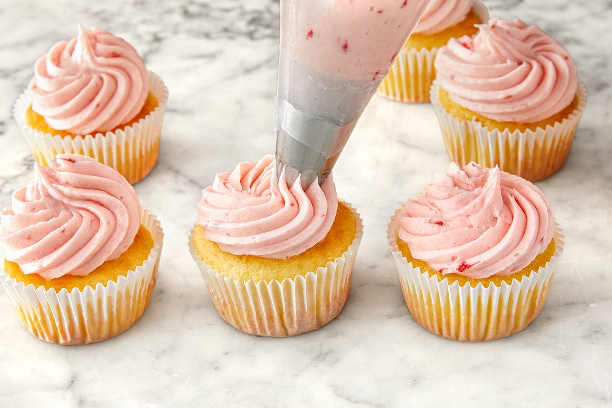 A piping bag with pink frosting is being used to decorate a yellow cupcake. Five other cupcakes with pink swirled frosting are on a marble countertop.