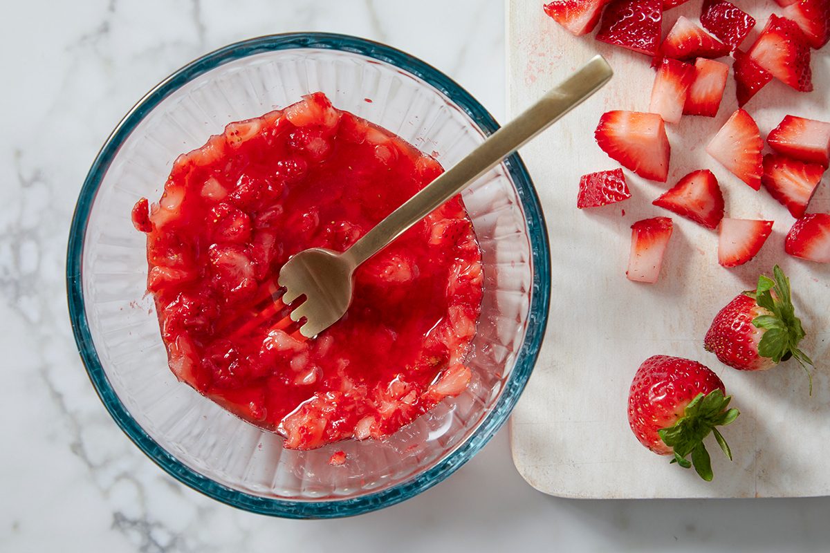A glass bowl filled with mashed strawberries and a small wooden fork on a marble surface. Next to it, a cutting board with diced and whole strawberries. Some strawberries have leaves attached.