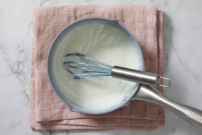 A saucepan with creamy white sauce and a whisk inside sits on a folded pink fabric on a marble surface.