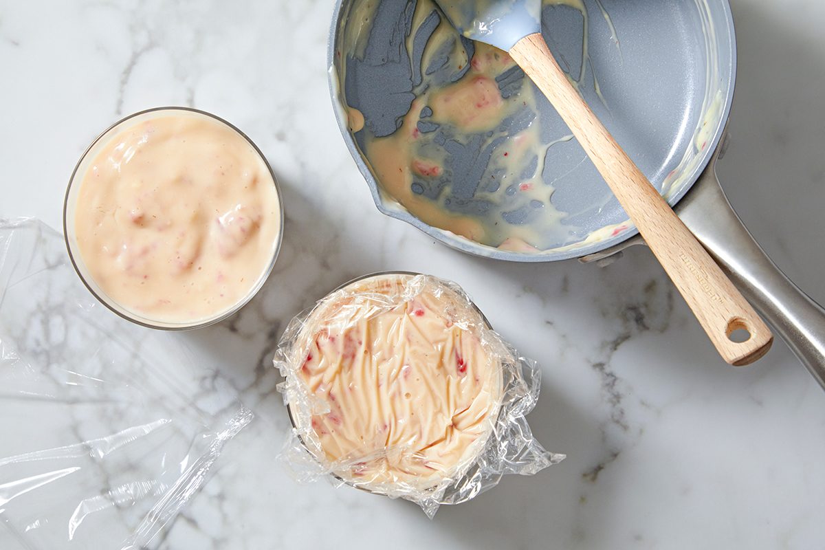 A bowl partially filled with a creamy mixture and a spatula beside two smaller containers. One container is filled with the mixture; the other is covered with plastic wrap. All items are on a marble surface.