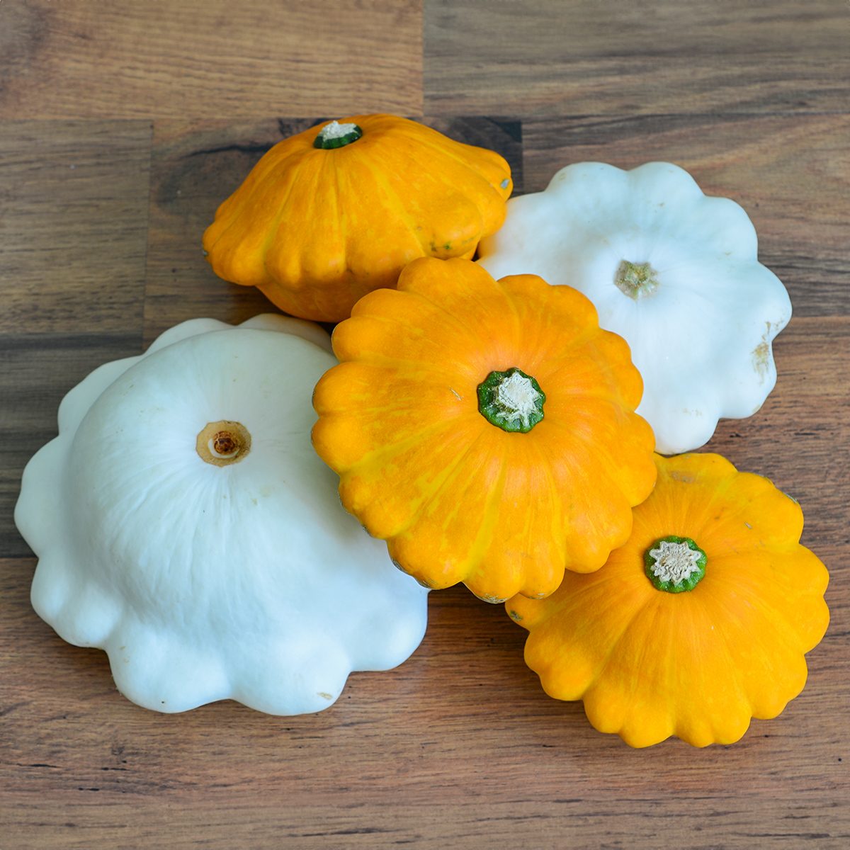 Five pattypan squashes, three yellow and two white, are arranged on a wooden surface. The squashes have scalloped edges and a central green stem.