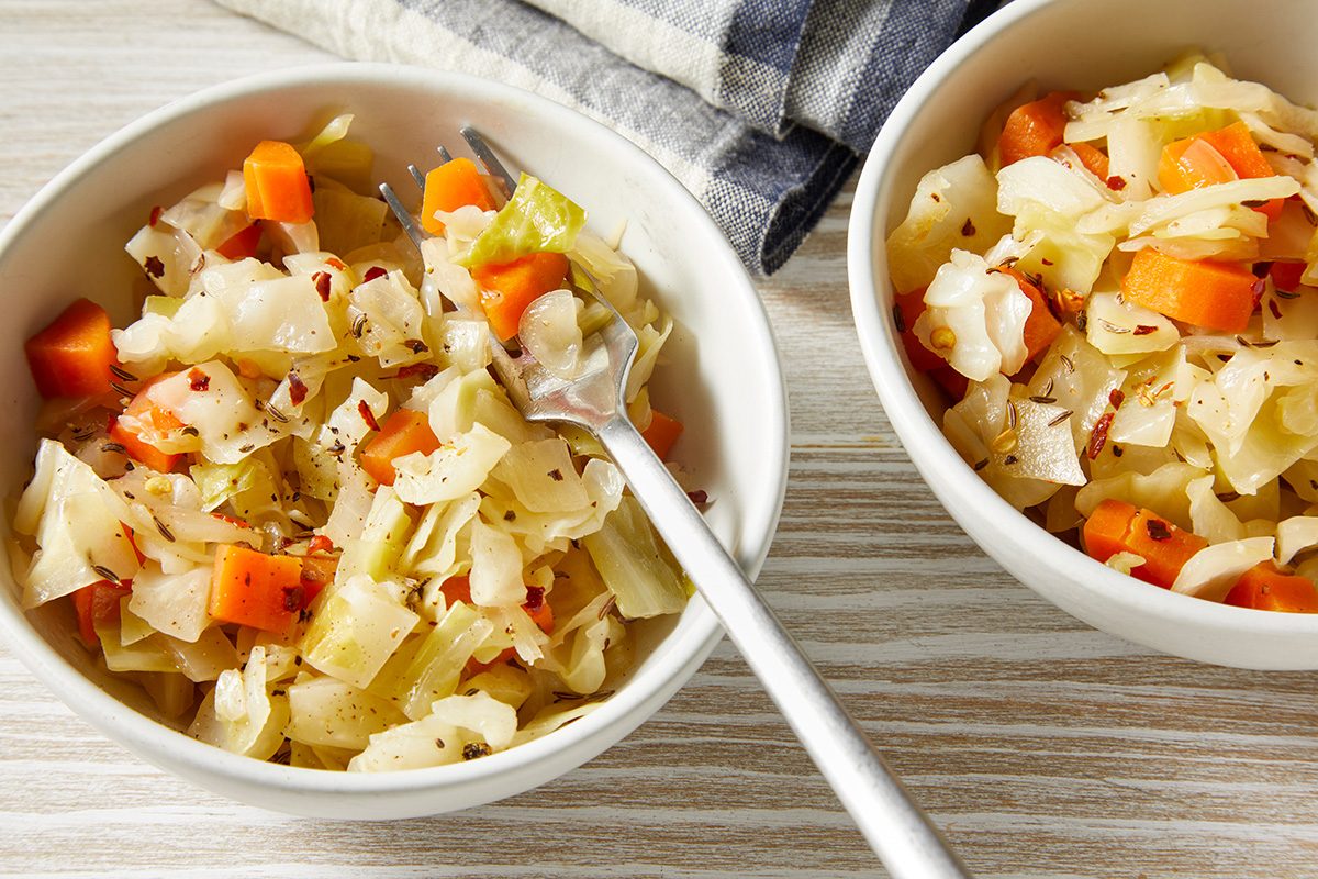 Two bowls of sautéed cabbage and carrots sit on a wooden surface. Each bowl contains chopped cabbage and carrot pieces, garnished with a sprinkle of red pepper flakes. A fork rests in one bowl, and a striped cloth is partially visible nearby.