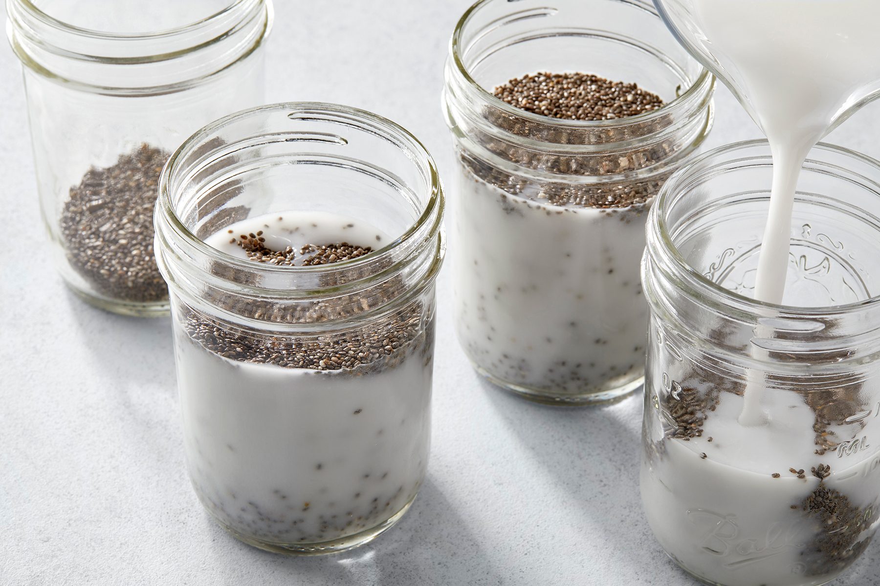 Four glass jars on a light surface, each containing chia seeds. Three jars are filled with a white liquid, and one jar is being filled as liquid is being poured in. The jars are arranged in a slightly scattered pattern.