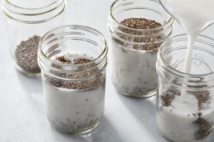 Four glass jars on a light surface, each containing chia seeds. Three jars are filled with a white liquid, and one jar is being filled as liquid is being poured in. The jars are arranged in a slightly scattered pattern.