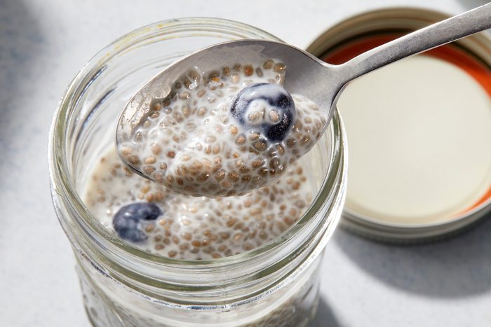 A glass jar filled with chia seed pudding and blueberries, with a spoon lifting out a portion. The jar's lid is partially visible beside it, and the background is softly blurred.