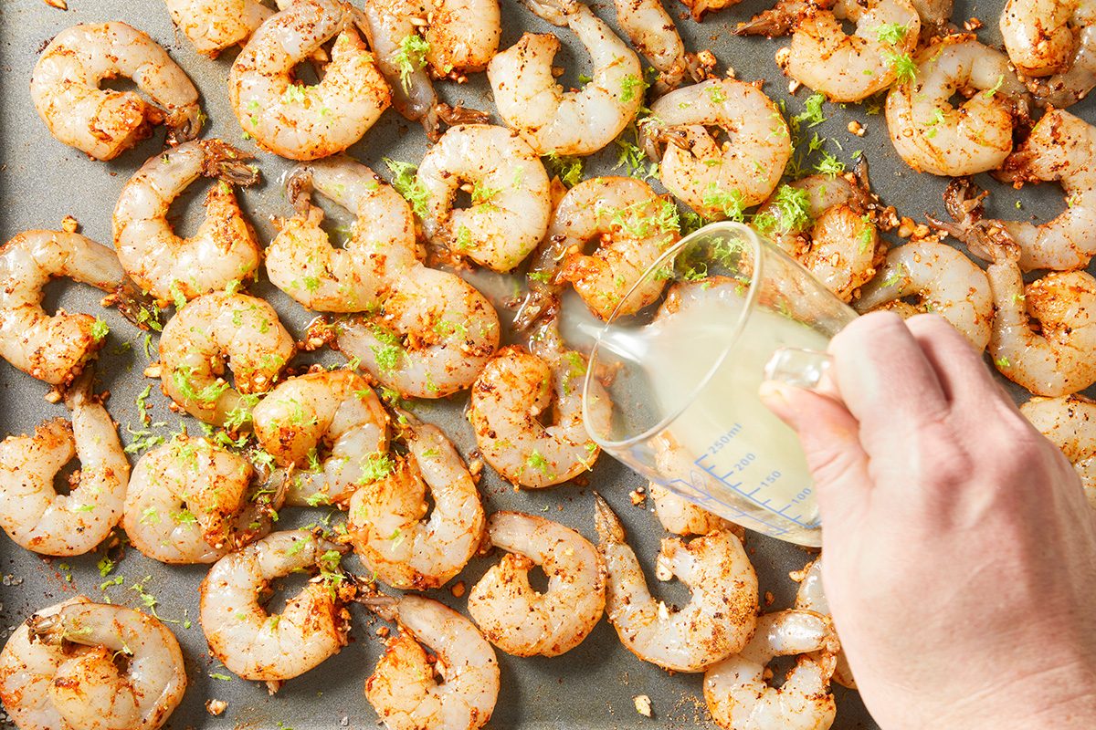 A hand pours lemon juice from a measuring cup over a baking sheet of raw, seasoned shrimp garnished with lime zest.