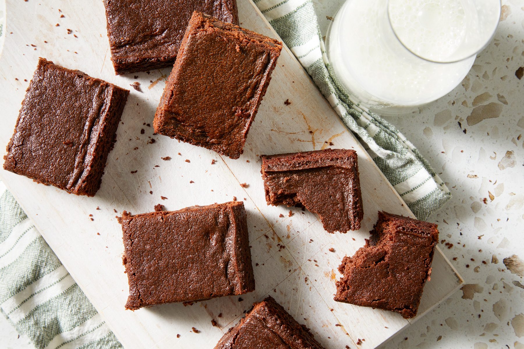 Overhead shot of Eggless Brownies; cut into pieces and served on white wooden tray over a napkin; A glass of milk is nearby; all set on a marble surface;