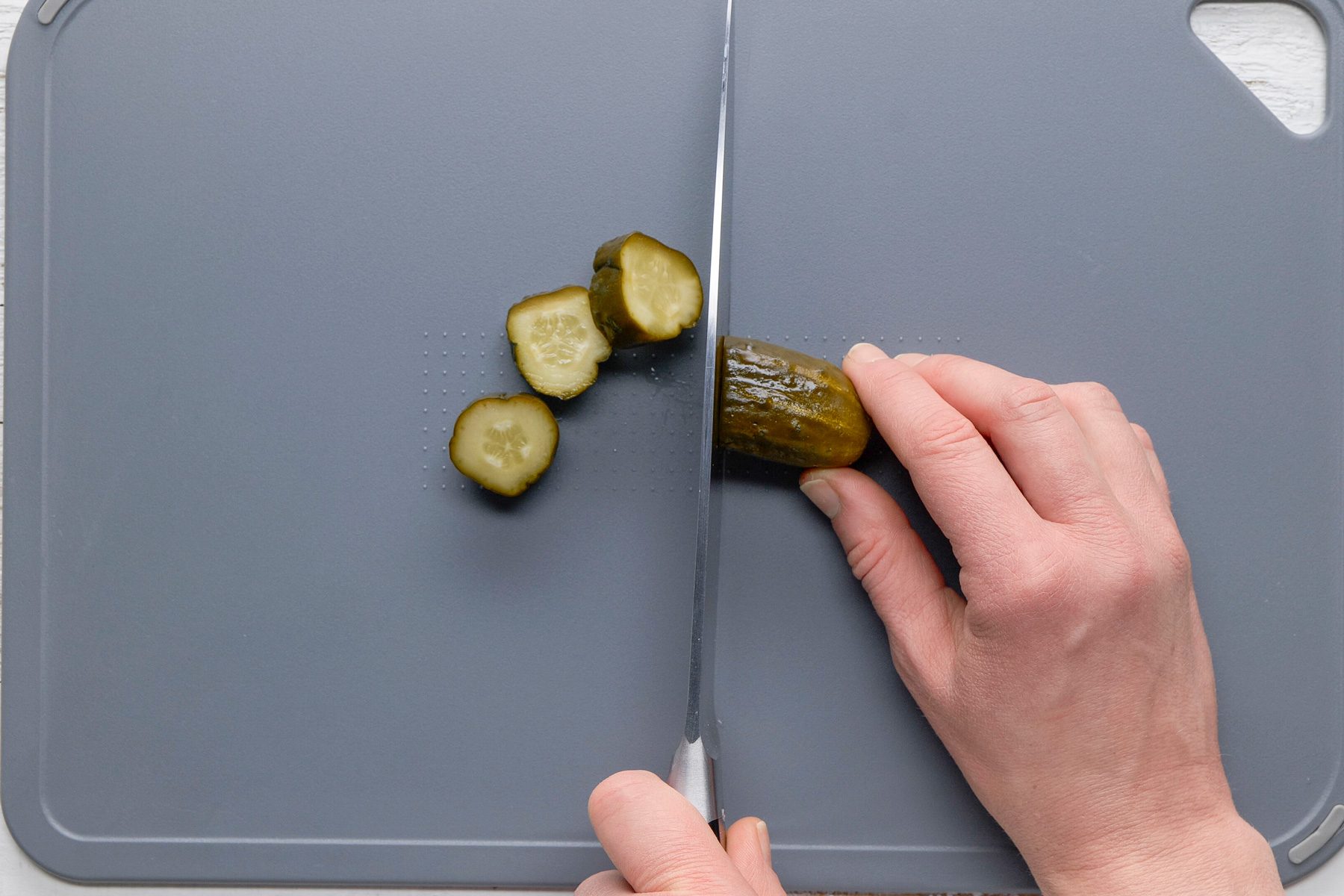 Overhead shot of drain pickles; discarding liquid; cut pickles into 1/2-inch-thick slices; chopping board; knife; white wooden surface;