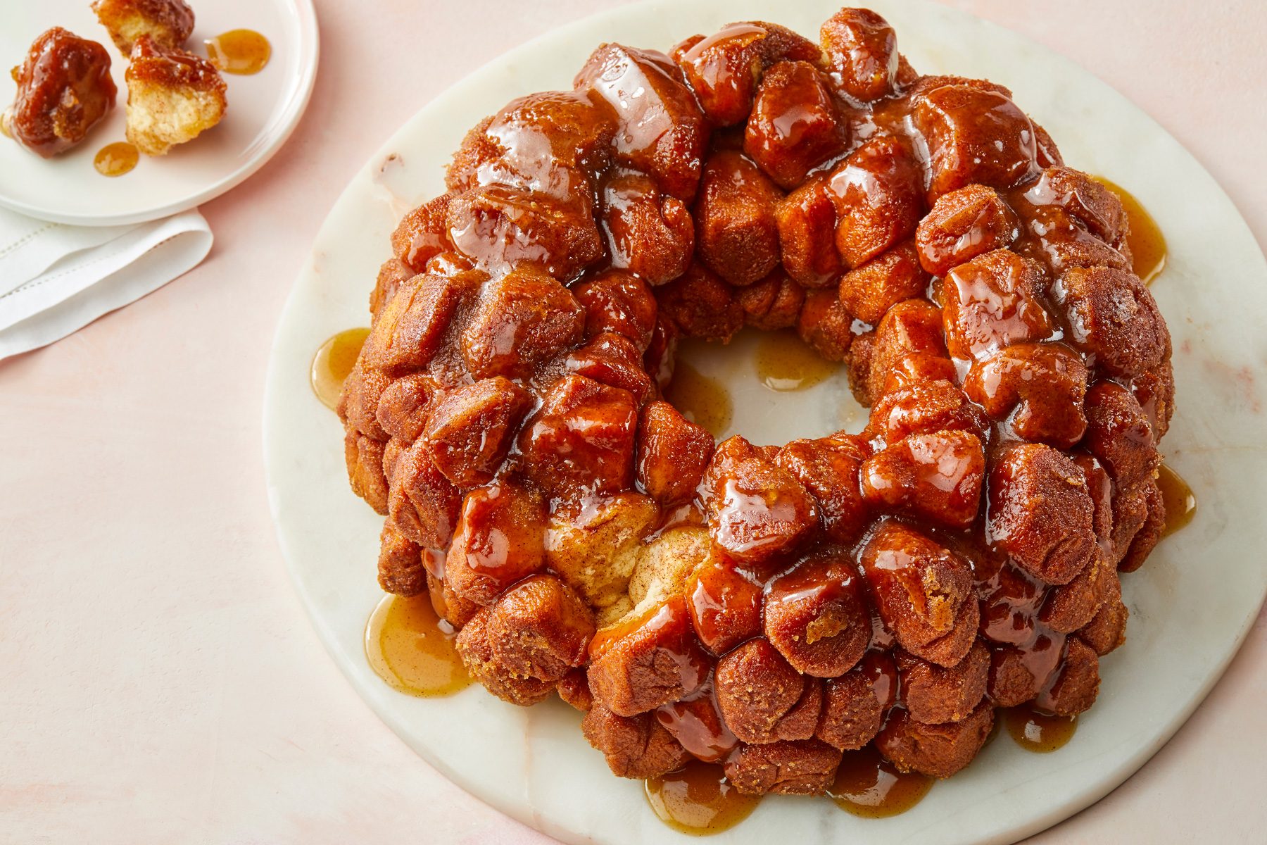3/4 angle view shot of Cinnamon Monkey Bread; Place on a round marble tray; served small piece on a small white plate; over a white napkin; all set on a light pink surface;