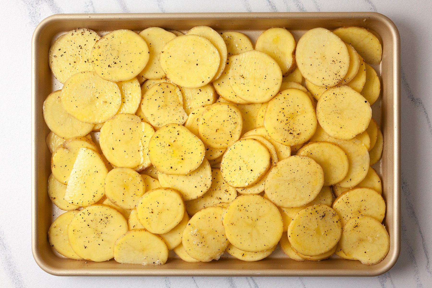 Sliced potatoes seasoned with black pepper and salt are arranged in a single layer on a baking sheet, ready for cooking.
