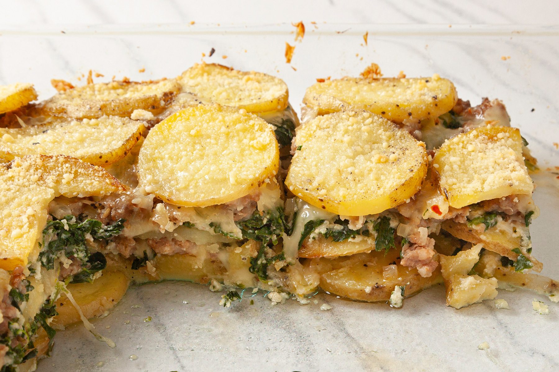 A close-up of a layered dish featuring thin potato slices, browned on top, with visible layers of cheese, spinach, and ground meat. The surface is sprinkled with grated cheese and black pepper.