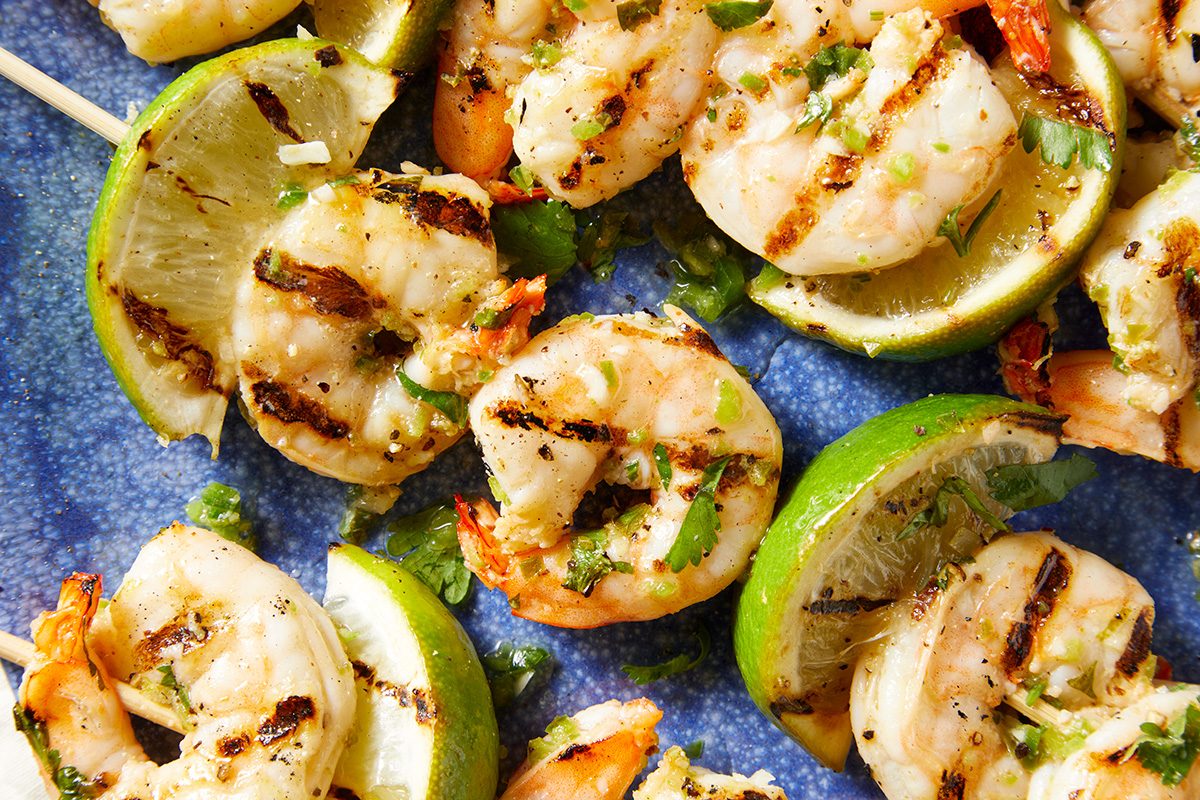 Grilled shrimp skewers with lime wedges arranged on a blue plate. The shrimp are seasoned with herbs and char marks are visible.