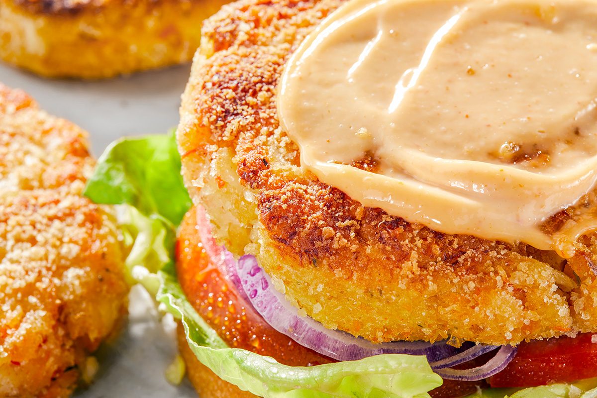 A close-up of a veggie burger with a crispy patty, fresh lettuce, sliced tomato, red onion, and a swirl of creamy sauce on top.