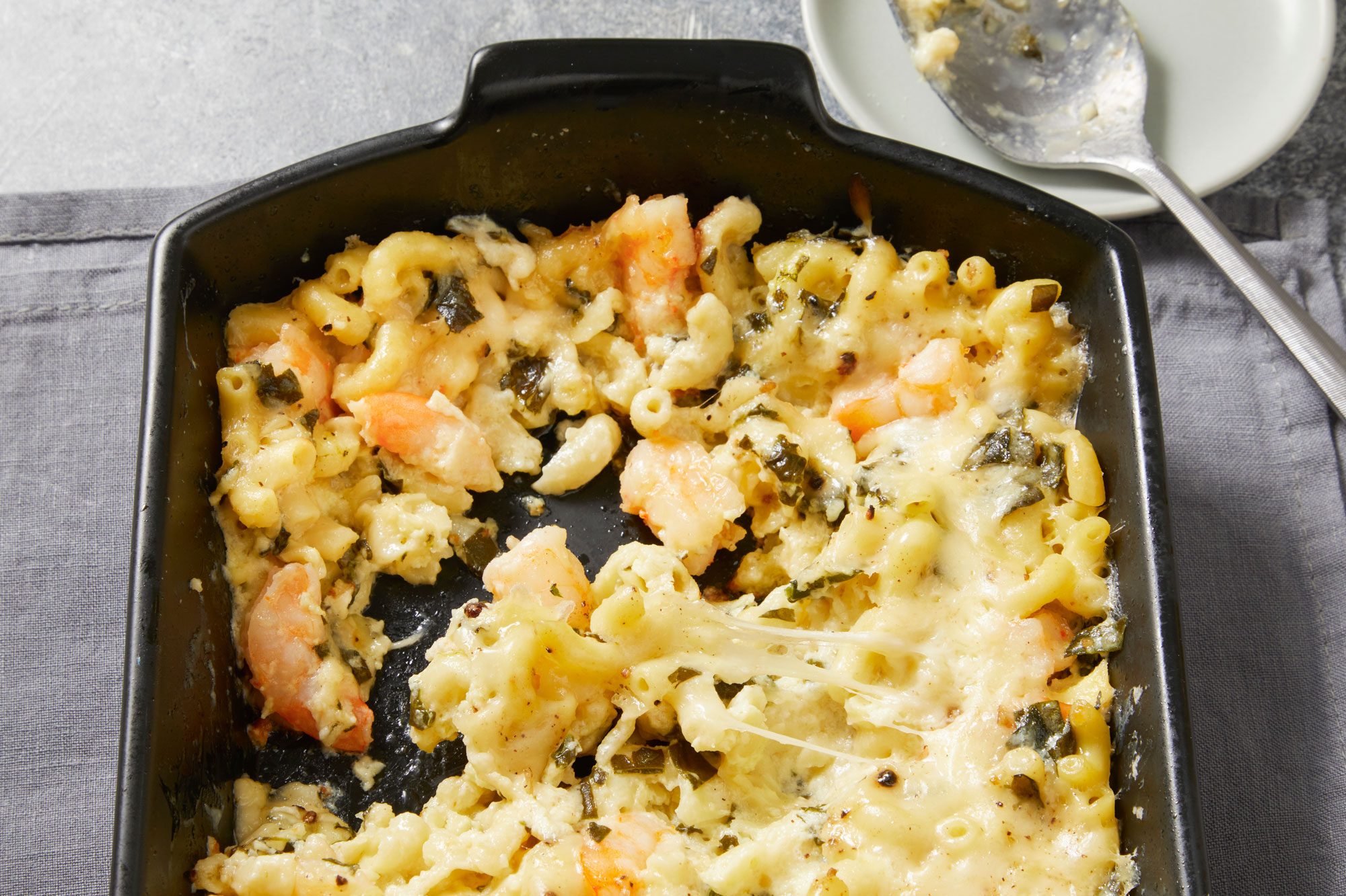 Close-up shot of Shrimp & Macaroni Casserole in a black baking pan placed on a grey napkin; a serving spoon rests on a small white plate to the right; set on a grey surface;
