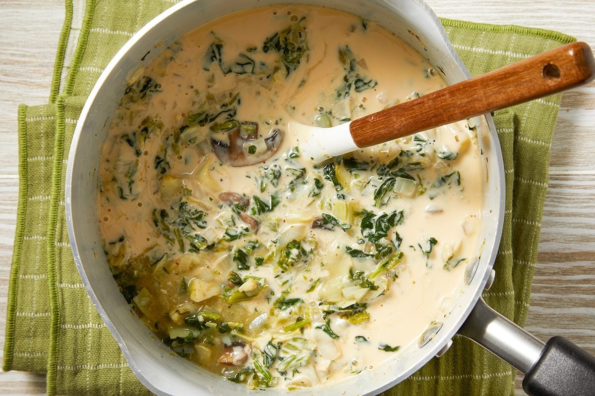 A pot of creamy soup containing leafy greens, set on a green cloth. A wooden-handled ladle rests inside, partially submerged in the soup, which appears to contain cheese or a similar ingredient.
