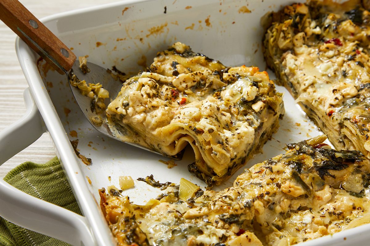A baked lasagna dish with a piece being lifted by a spatula. The lasagna is layered with pasta, cheese, and spinach, and is sprinkled with herbs and spices. It is in a white casserole dish with a green cloth underneath.