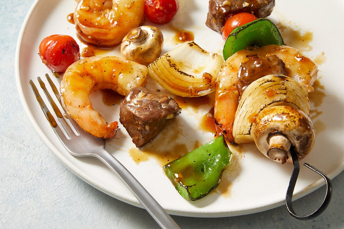 Grilled skewers on a white plate with shrimp, beef, mushrooms, onions, cherry tomatoes, and green bell peppers. A fork rests on the plate's edge.