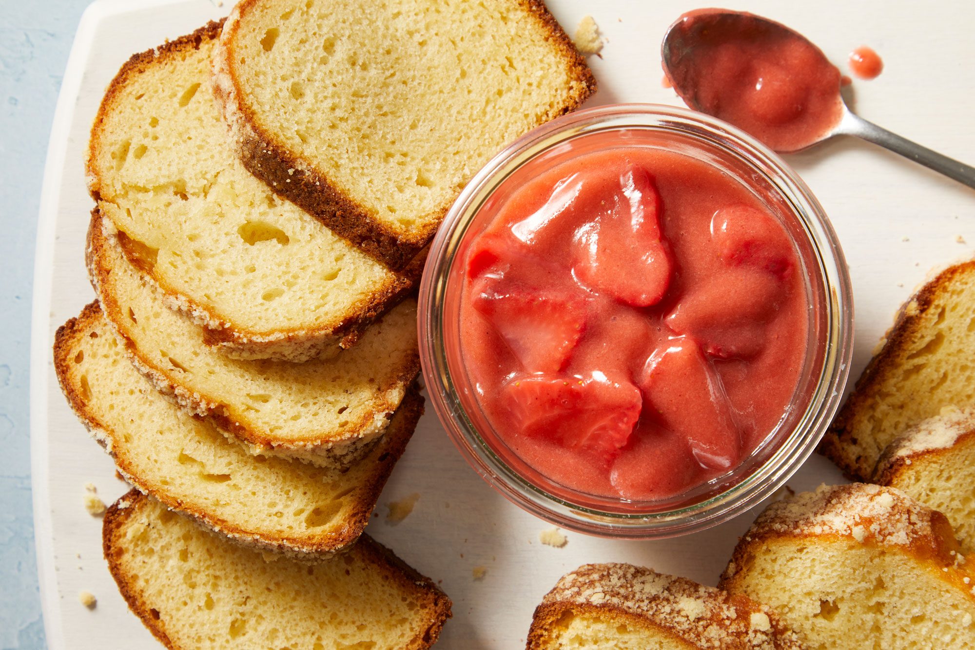 Overhead shot of Strawberry Rhubarb Sauce; served in a glass bowl with breads and a spoon on a white plate; all set up on a light blue surface;