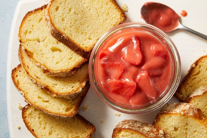 Overhead shot of Strawberry Rhubarb Sauce; served in a glass bowl with breads and a spoon on a white plate; all set up on a light blue surface;