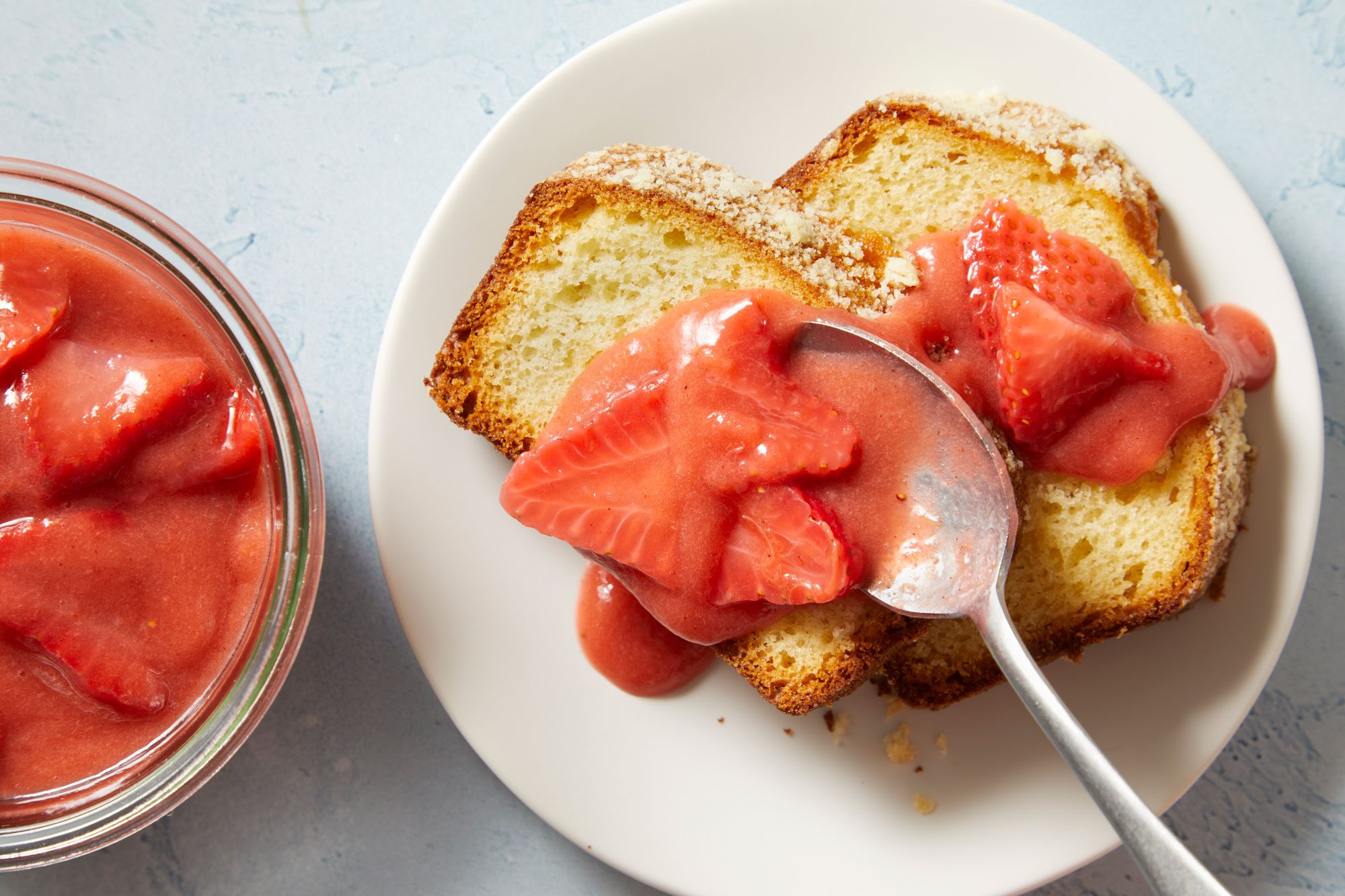 Overhead shot of Strawberry Rhubarb Sauce; in a glass bowl; served over breads on a white plate with a spoon; placed on a light blue surface