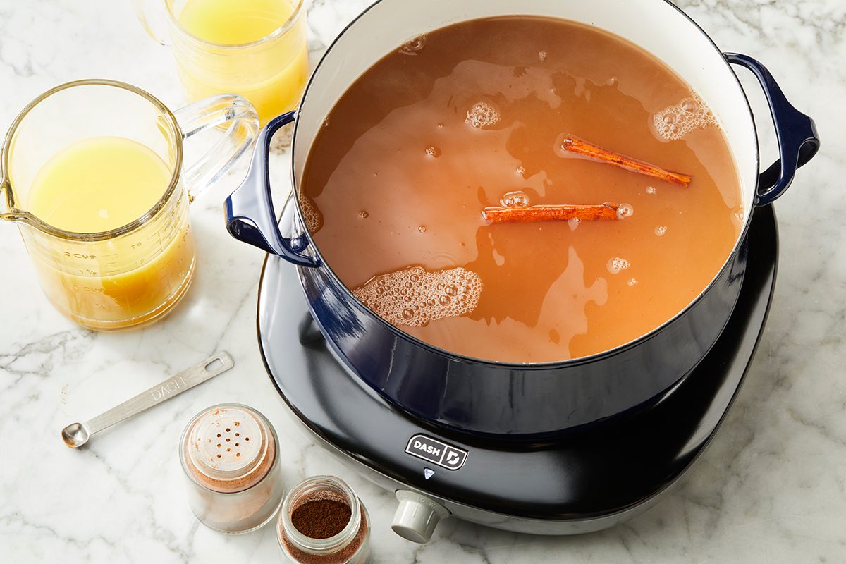 A blue pot on an electric burner contains a brown liquid with cinnamon sticks. Nearby are a measuring spoon, a glass pitcher of juice, and two small spice jars on a marble countertop.