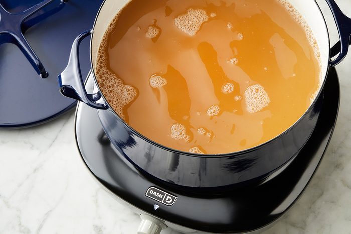 A blue pot filled with simmering light brown broth sits on an electric burner, with its lid resting nearby on a white marble countertop.