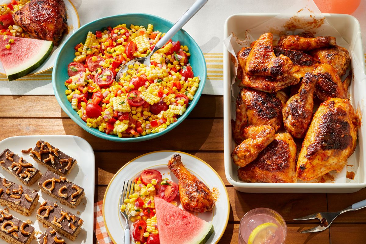 3/4th shot of a vibrant outdoor dining scene with a variety of delicious dishes placed over wooden background, A platter of Barbecued Chicken takes center stage, featuring crispy, golden-brown chicken pieces coated in a tangy barbecue sauce, A bowl of Sweet Corn Tomato Salad, A refreshing watermelon slice and a glass of lemonade complement the meal, while a tray of bite-sized desserts completes the feast,The setting, bathed in natural light, conveys a summery, inviting atmosphere perfect for a shared meal;