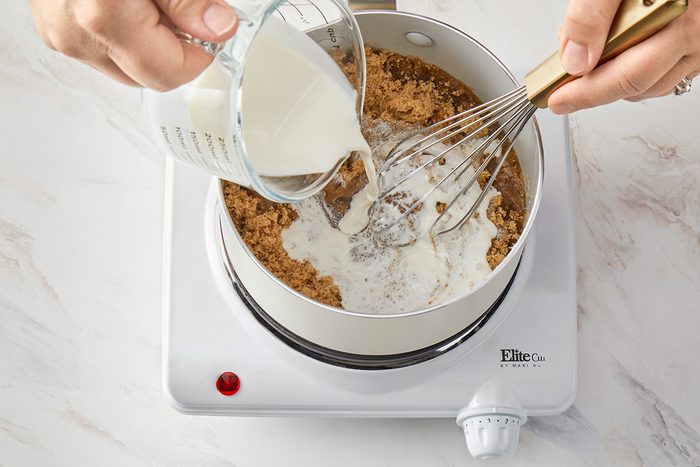 3/4th shot of a person pours milk from a measuring cup into a pot with brown sugar while whisking the mixture on a white electric stove; the background is a white textured marble countertop