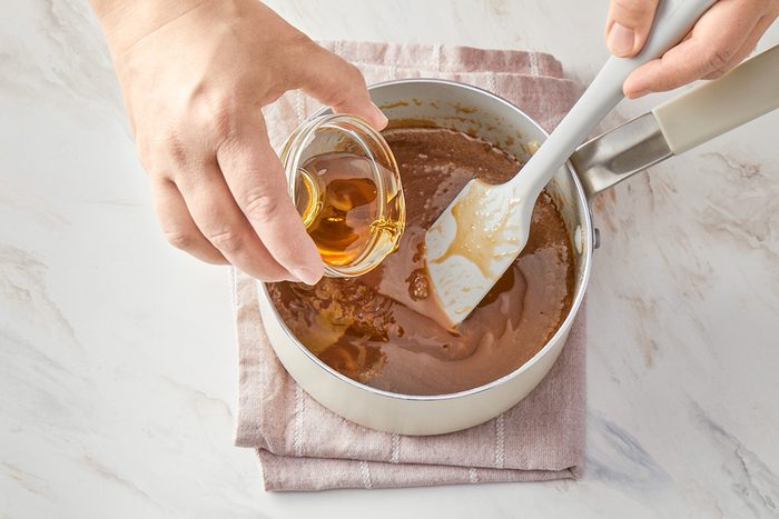 3/4th shot of A person pours a small glass bowl of vanilla extract, into a saucepan containing a light brown mixture, stirring with a spatula, The pan rests on a folded cloth on a marble surface