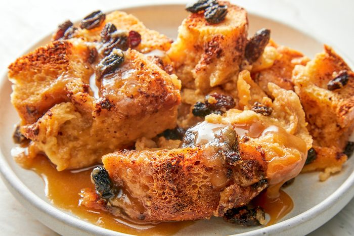 closeup shot of a plate of Bread Pudding With Rum Sauce topped with raisins and drizzled with caramel sauce, served on a light colored dish