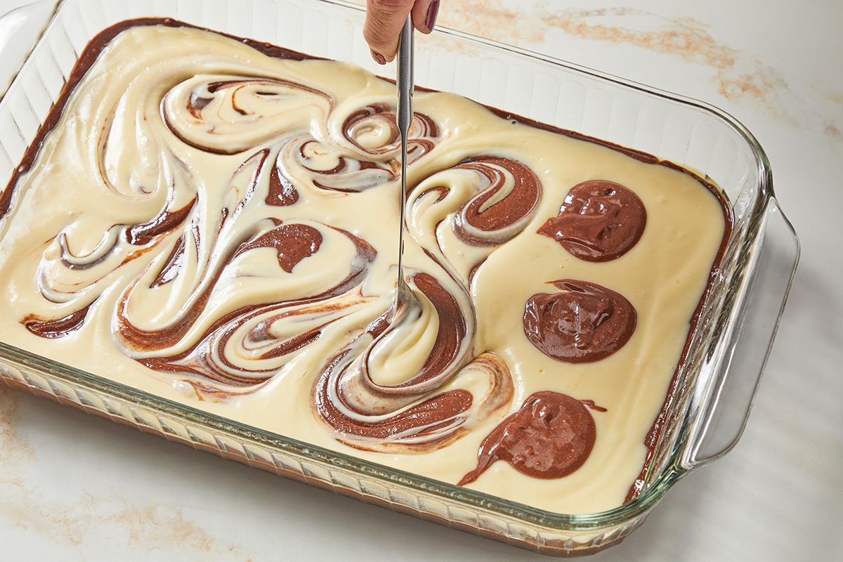 3/4 angle view shot of a glass baking dish; Cut through batter with a knife to swirl; Bake at 350 degree for 28-32 minutes or until a toothpick inserted in the center comes out with moist crumbs (brownies may appear moist); Cool completely on a wire rack; Spread frosting over brownies; all set on a marble surface