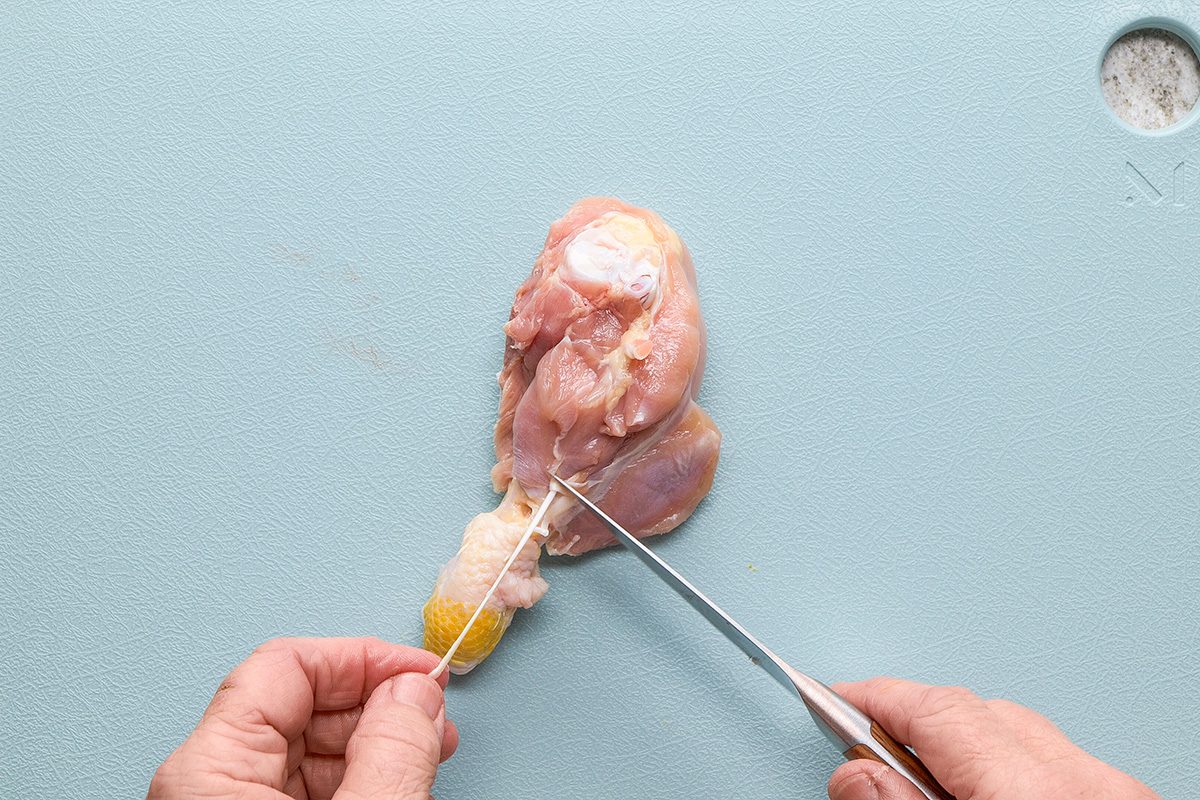 A person uses a knife and kitchen twine to prepare a raw chicken drumstick on a light blue cutting board.