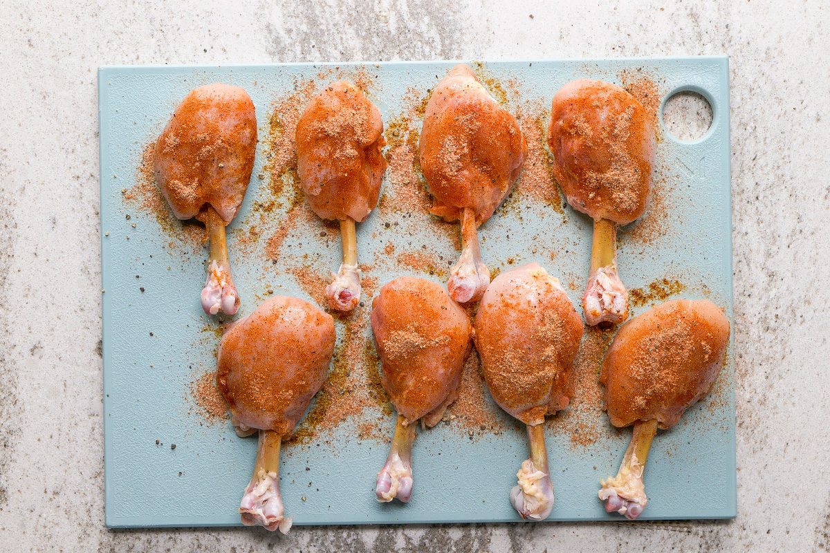Eight raw chicken drumsticks coated with seasoning are arranged on a light blue cutting board, which is placed on a light-colored countertop.