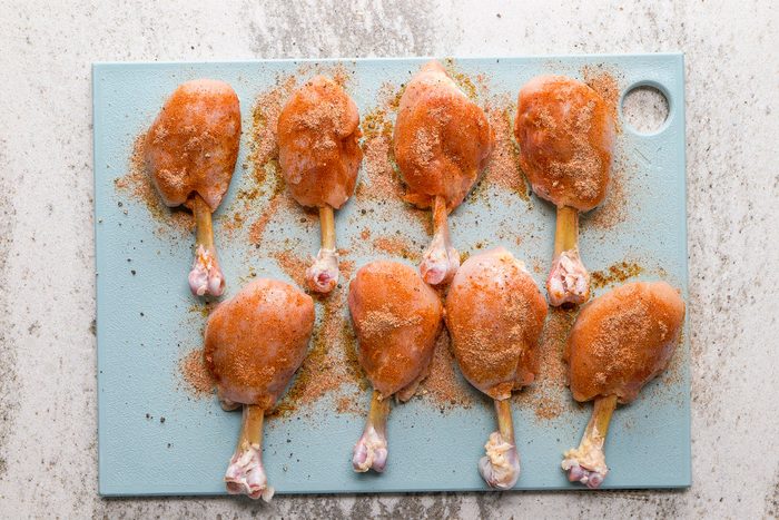 Eight raw chicken drumsticks coated with seasoning are arranged on a light blue cutting board, which is placed on a light-colored countertop.