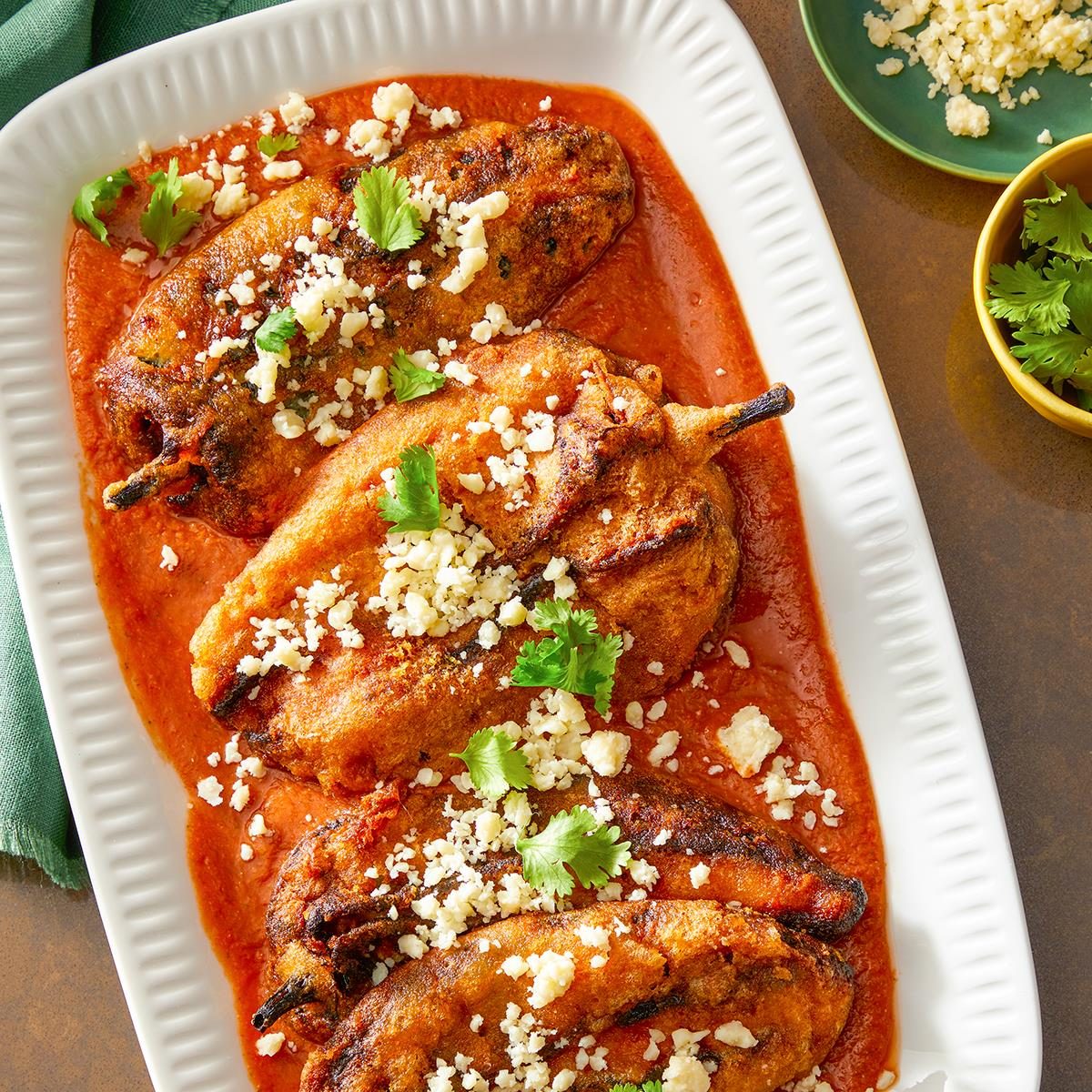 Top view shot of Chile Rellenos; To serve spoon red sauce in shallow bowls; top with fried peppers; Sprinkle with cilantro and Cotija cheese; Serve immediately; all arranged on a light brown surface