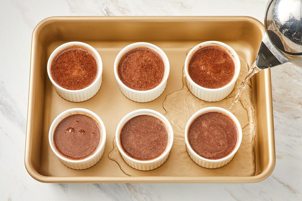 Six ramekins filled with chocolate custard are placed in a gold baking pan, while hot water is being poured into the pan around the ramekins.