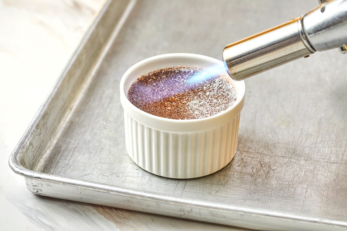 A small white ramekin on a metal baking sheet is being caramelized with a kitchen torch, browning the sugar on top of a crème brûlée.