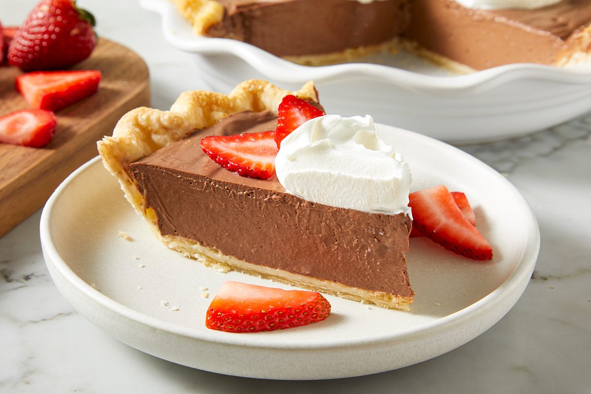 A slice of chocolate pie topped with whipped cream and sliced strawberries sits on a white plate, with more strawberries and pie in the background.