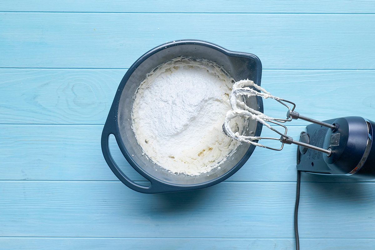 Overhead shot of gradually beat in enough confectioners' sugar to achieve desired consistency; Store in the refrigerator; blender; blue wooden surface