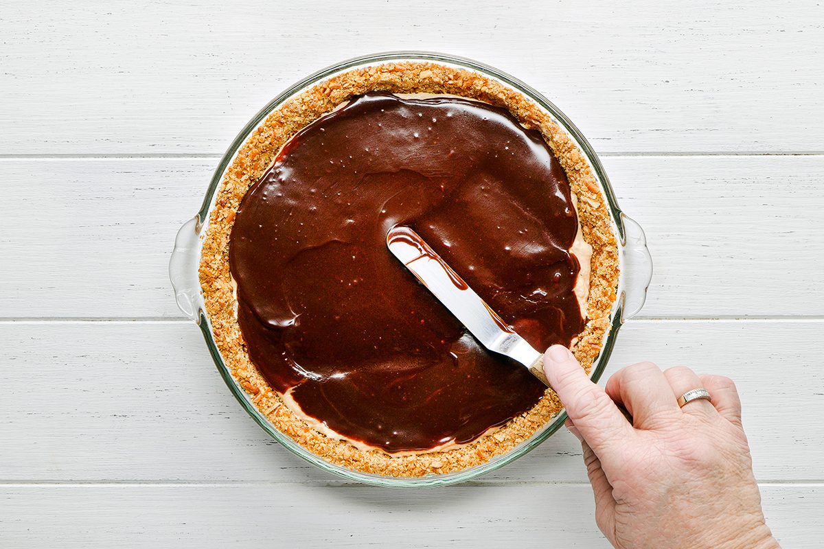 A hand spreading a smooth layer of chocolate filling over a pie crust with an offset spatula on a white wooden surface.