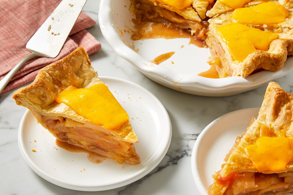A slice of apple pie topped with melted cheddar cheese sits on a white plate, with more pie and slices in a ceramic dish nearby on a marble surface. A pie server and pink cloth are in the background.