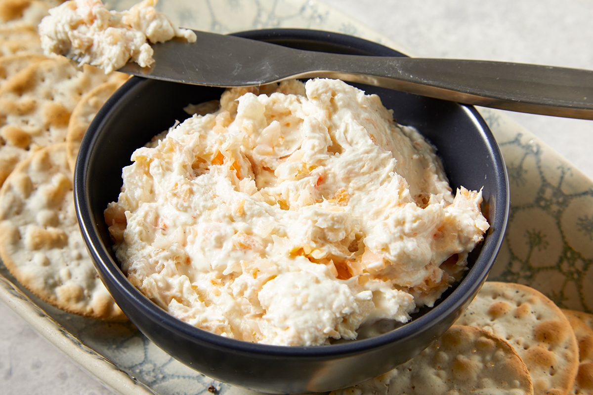A bowl of creamy cheese spread with a knife resting on top, surrounded by round crackers on a patterned tray.